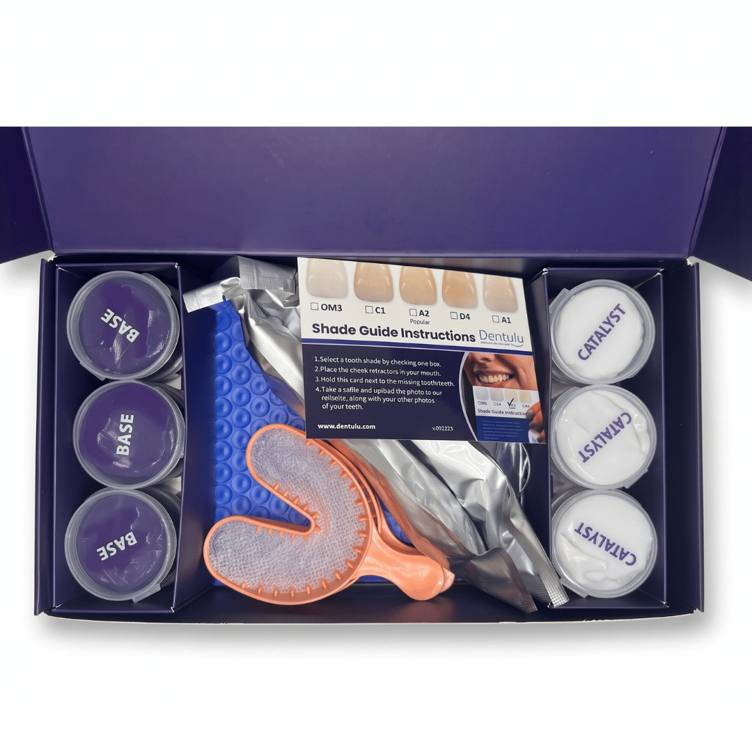 An open dark purple box containing a dental shade guide kit with two small round jars labeled BASE and CATALYST, a shade guide card, a blue textured tray, an orange-handled brush, and other dental accessories arranged in a tray insert.