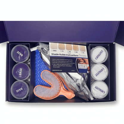 An open dark purple box containing a dental shade guide kit with two small round jars labeled BASE and CATALYST, a shade guide card, a blue textured tray, an orange-handled brush, and other dental accessories arranged in a tray insert.